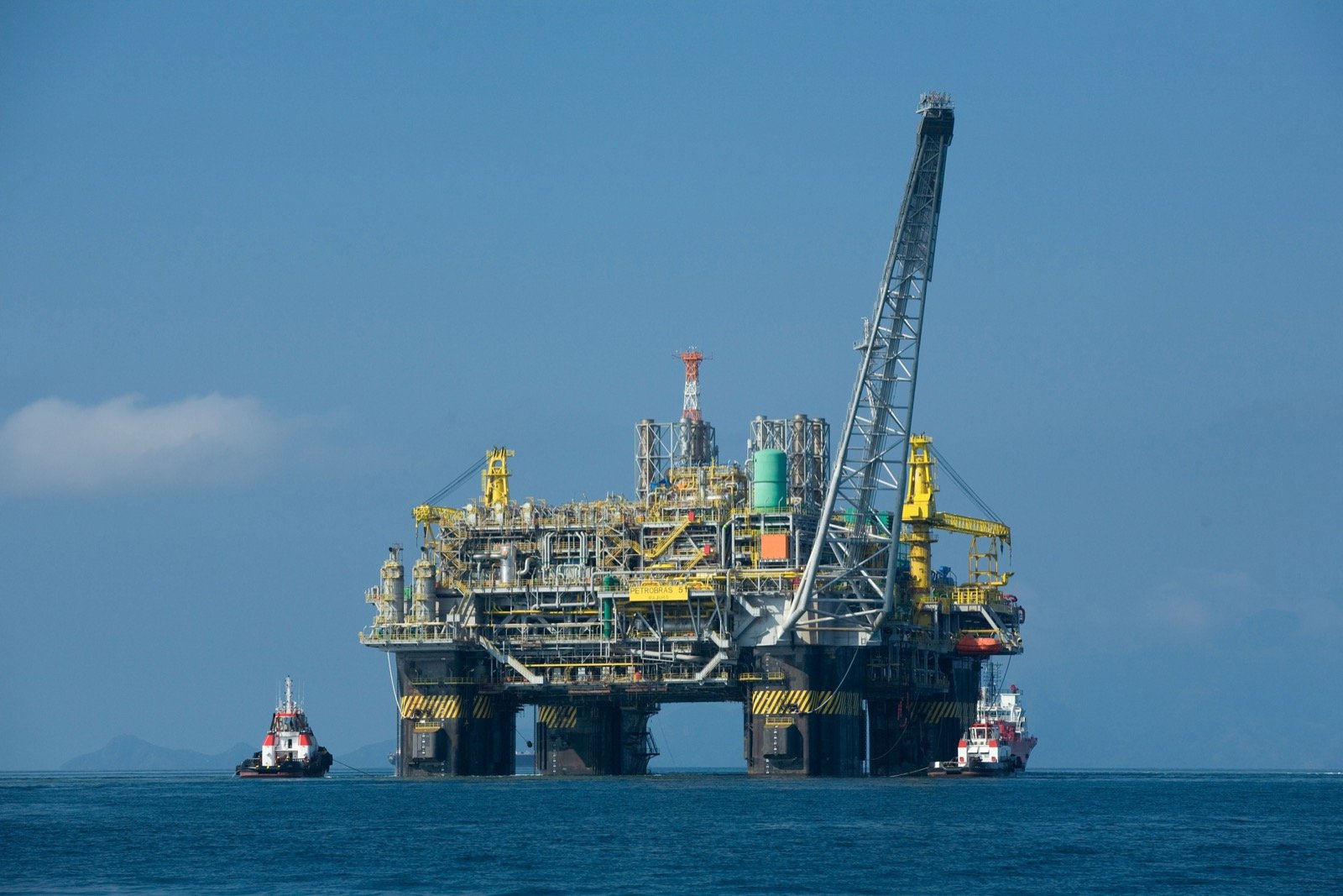 Plataforma de petróleo semissubmersível amarela e cinza no mar, com dois rebocadores ao lado