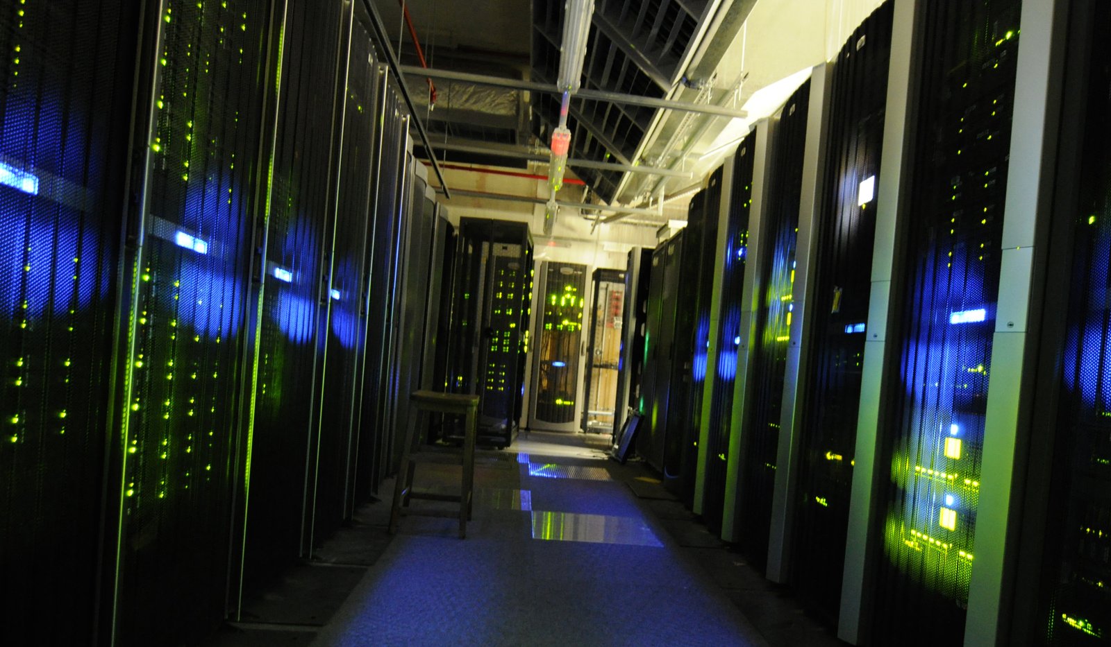 Corredor escuro de uma sala de servidores, com armários (racks) dos dois lados e luzes verdes e azuis acesas.