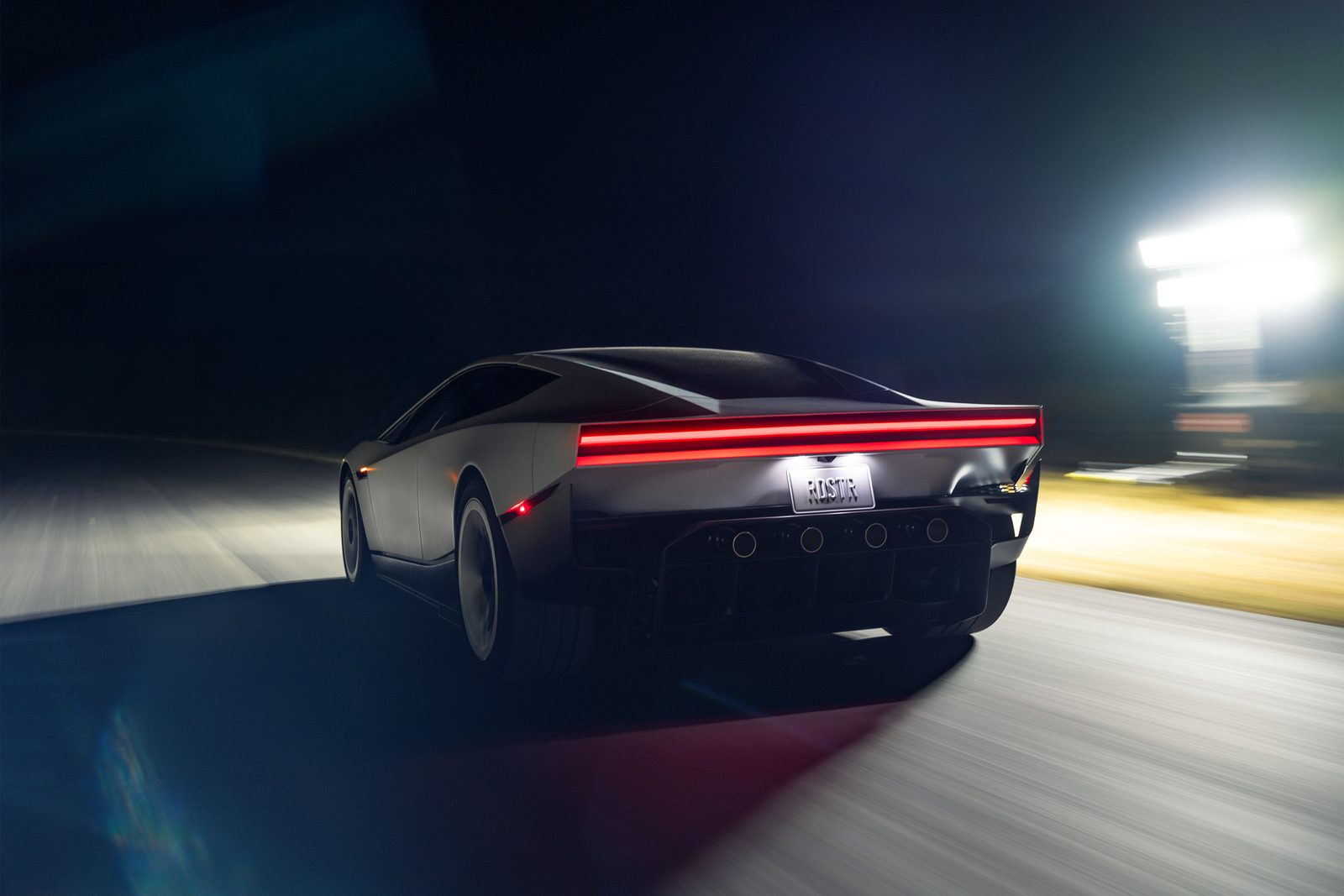 Carro esportivo prateado visto de trás em movimento numa via à noite, com faixa de luz vermelha na traseira