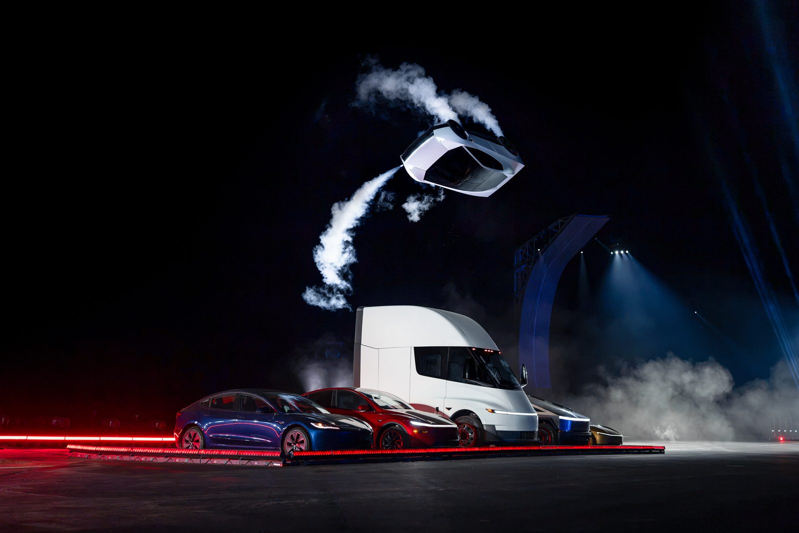 Carro esportivo branco no ar, deixando rastro de fumaça, sobre uma fila de carros Tesla e um caminhão Semi branco, em palco escuro